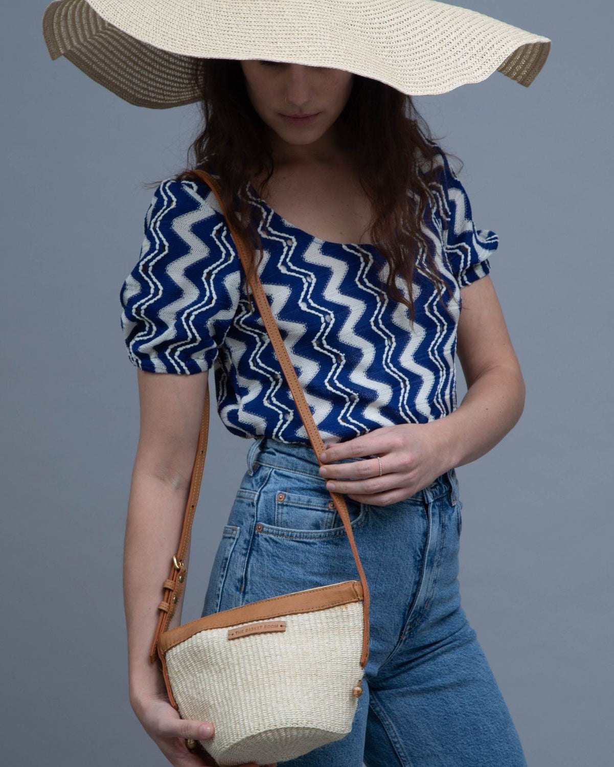 A woman wearing a wide-brimmed straw hat, a blue and white zigzag patterned top, and blue jeans carries a woven straw crossbody bag with tan leather trim against a solid gray studio background.