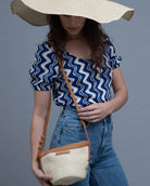 A woman wearing a wide-brimmed straw hat, a blue and white zigzag patterned top, and blue jeans carries a woven straw crossbody bag with tan leather trim against a solid gray studio background.