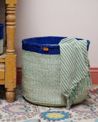 Woven basket with a blue top and green base, draped with a green and white striped towel on a patterned rug.