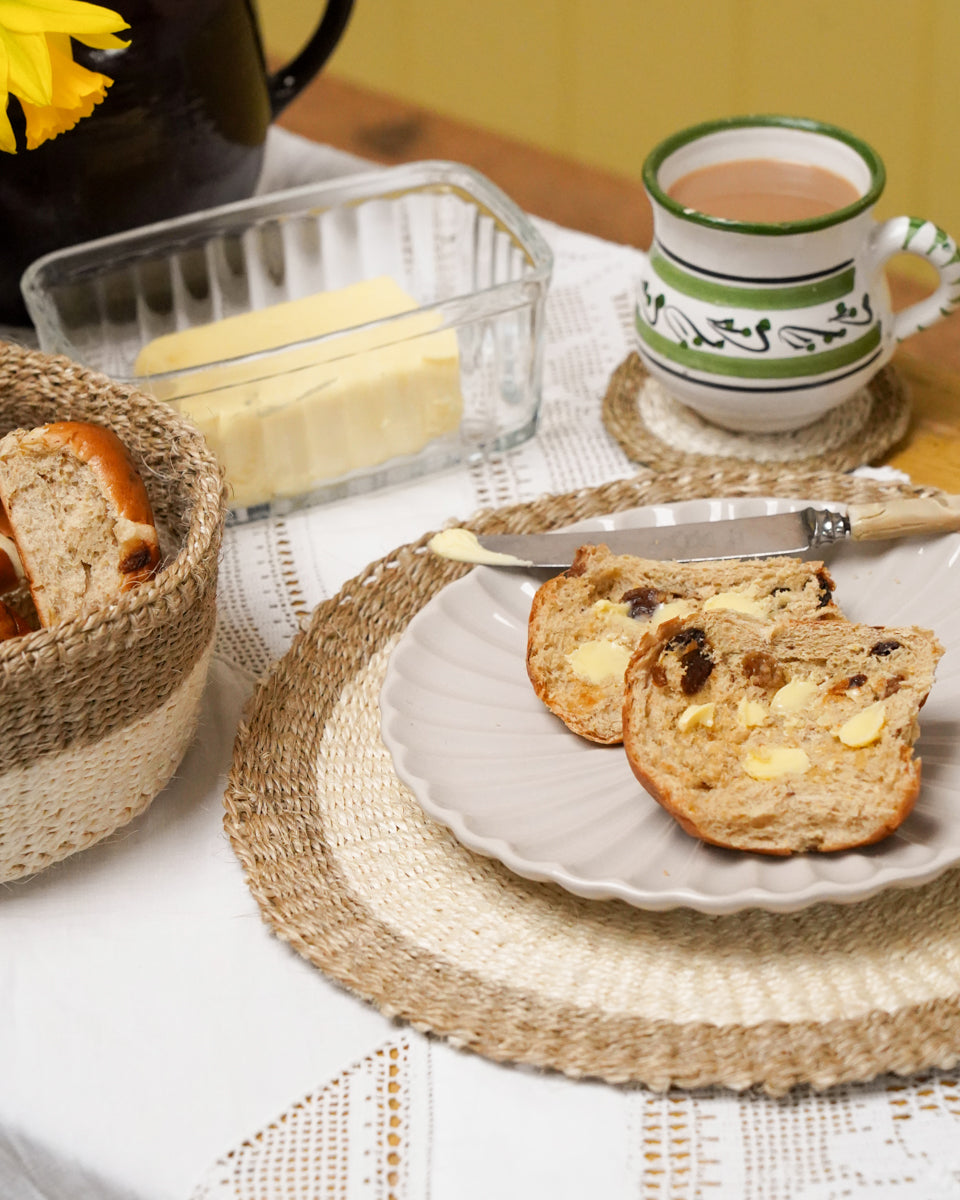 Natural woven placemat, coaster and bread basket on a table with a tea pot and hot cross bun