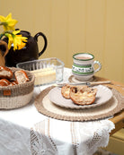 Tea time setting with bread, tea cup, and teapot on a table. Woven placemat, bread basket and coaster.