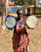 Woman holding two woven placemats outdoors with a rural background