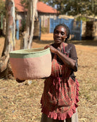 Woman holding a colorful woven basket in an outdoor setting with rustic buildings and trees.