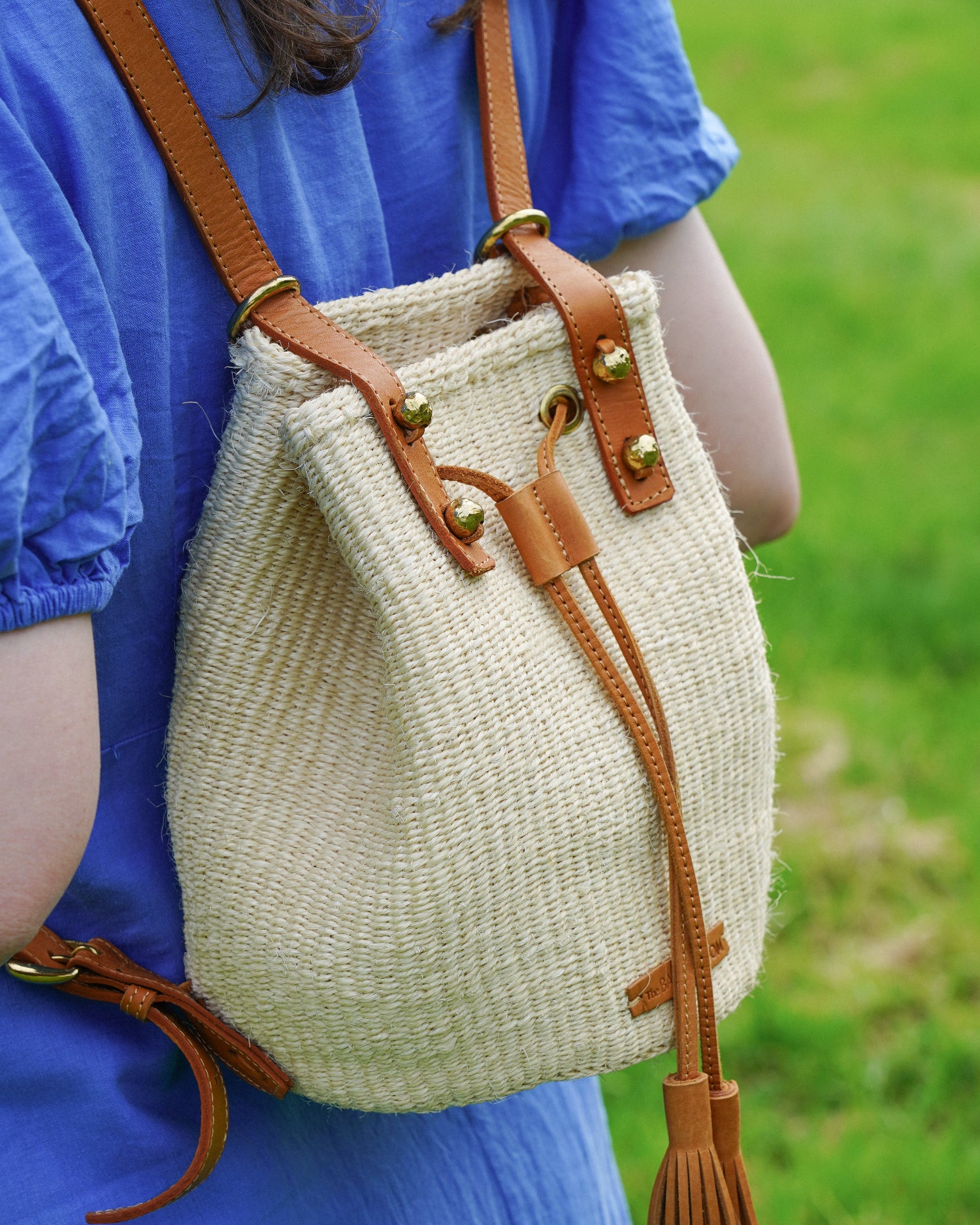 A woven straw bucket bag with tan leather straps and gold-tone hardware, shown worn on a person in a blue dress against a green grassy background.