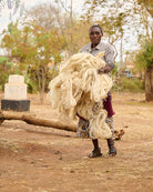 Basket weaver carrying a large bundle of sisal outdoors with trees and a building in the background.