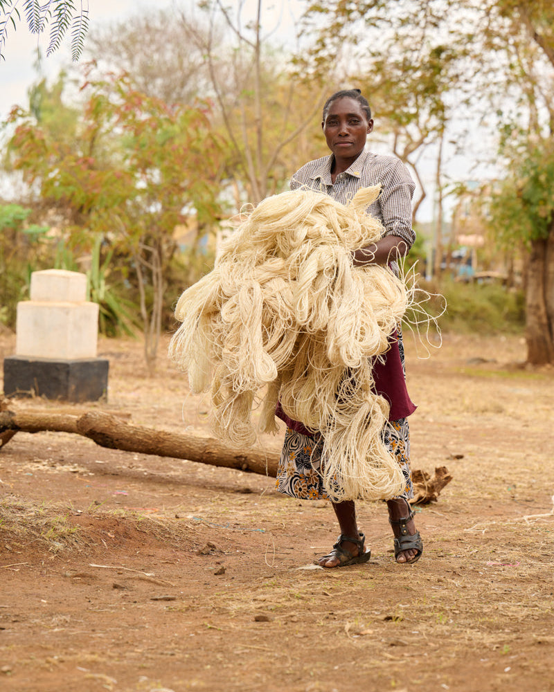 Basket weaver carrying a large bundle of sisal outdoors with trees and a building in the background.